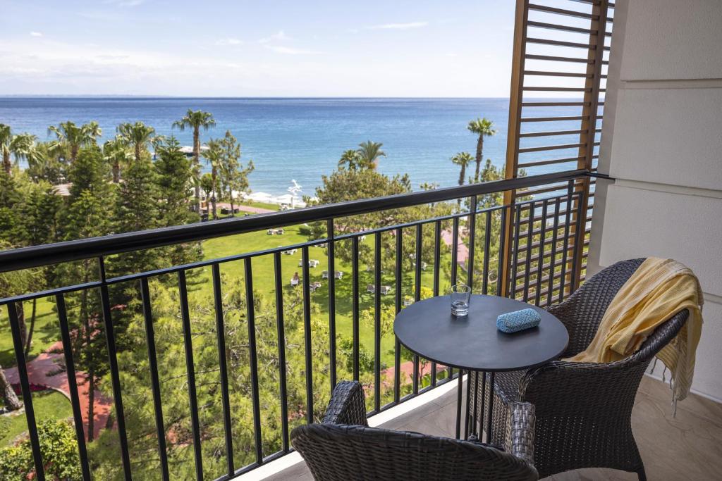 Skats no balkona uz jūru, kur redzamas palmu rindas un zaļš zāliens. Uz galda atrodas ūdens glāze un krāsa.