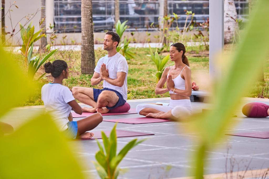 Grupas jogas nodarbība ārā, kur dalībnieki meditē un veic jogas pozas, ieskauti zaļumos un dabā.