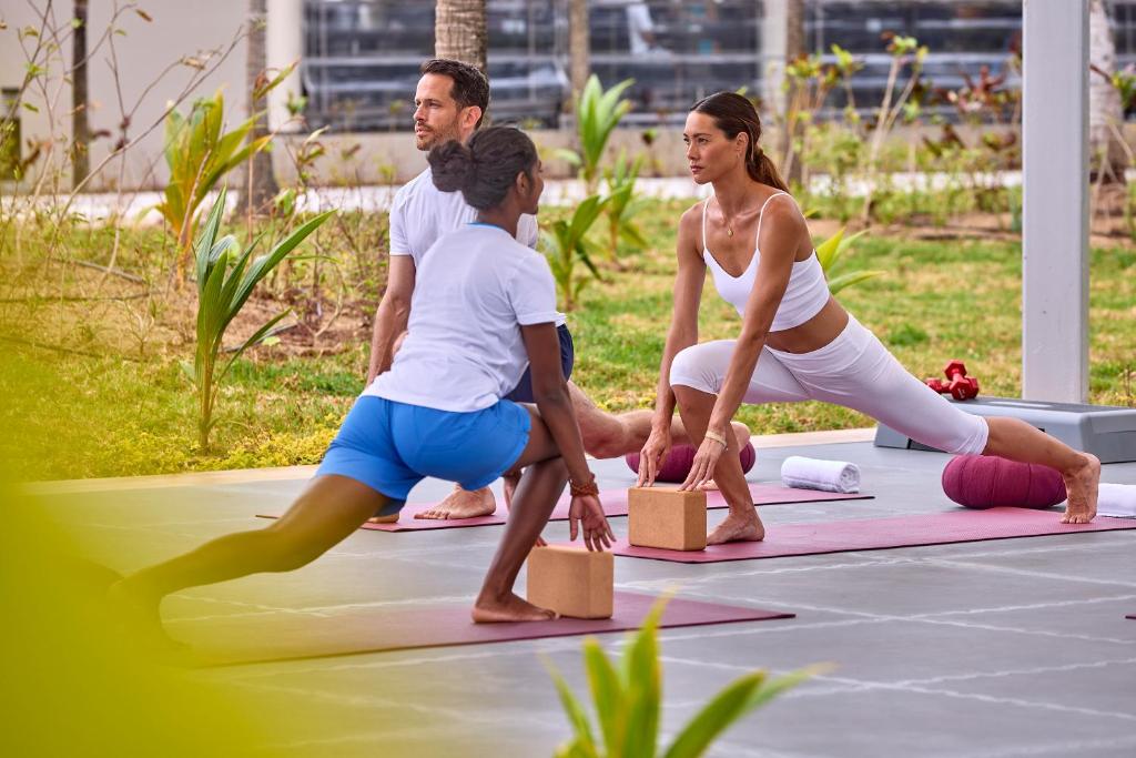 Joga nodarbība brīvā dabā ar trim dalībniekiem, kuri veic pozas uz paklājiem, izmantojot jogas blokus. Apkārt ir zaļumi un mīksts apgaismojums.