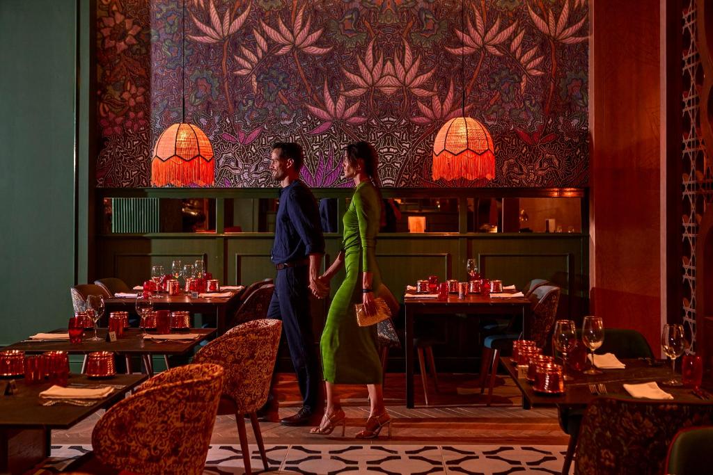 Eleganta pāra gājiens cauri stilīgai restorāna telpai ar krāsainu sienu dekoru un izsmalcinātiem gaismas ķermeņiem. Restorāns ir piepildīts ar gaumīgi iestatītiem galdiem un romantisku atmosfēru.