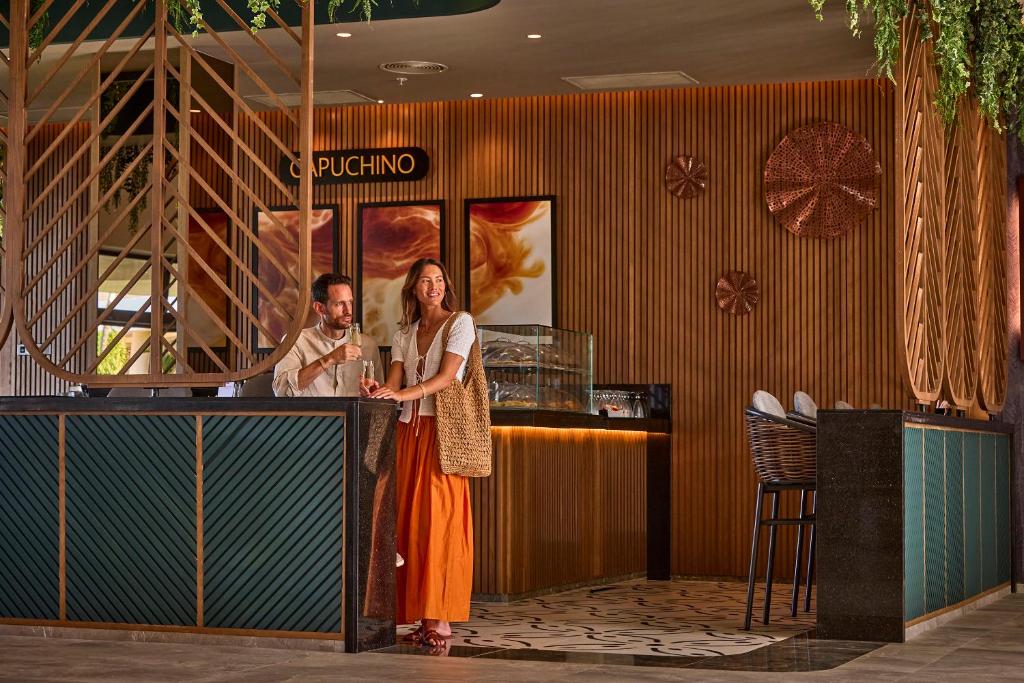 Kafijas bārs ar modernu interjeru, kur pāris izbauda dzērienus pie elegantas letes. Sienas rotā koka apdare un mākslas darbi, radot siltu atmosfēru.