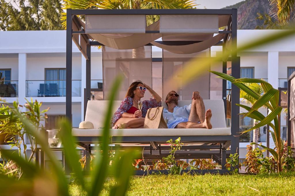 Pāris atpūšas modernas koka lapenē, baudot sauli un dabu. Apkārt ir zaļa zāle un palmām.