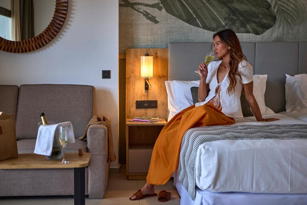 Sieviete sēž viesnīcas numurā, baudot kokteili, blakus ir šampanieša pudele un eleganta dekora elementi. Numurs ir gaišs un moderns, radot relaksējošu atmosfēru.