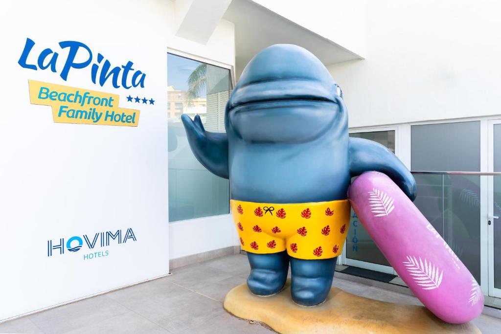 Lielizmēra zila figūra ar dzeltenām šortiem un rozā pludmales spilvenu, kas stāv pie "La Pinta" viesnīcas ieejas. Viesnīcas nosaukums un logo redzami uz sienas.
