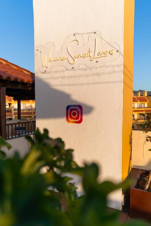 Alt tekst: Gaismas zīme "We are Sunset Lovers" uz baltas sienas ar Instagram logotipu, apzaļumotā vidē.
