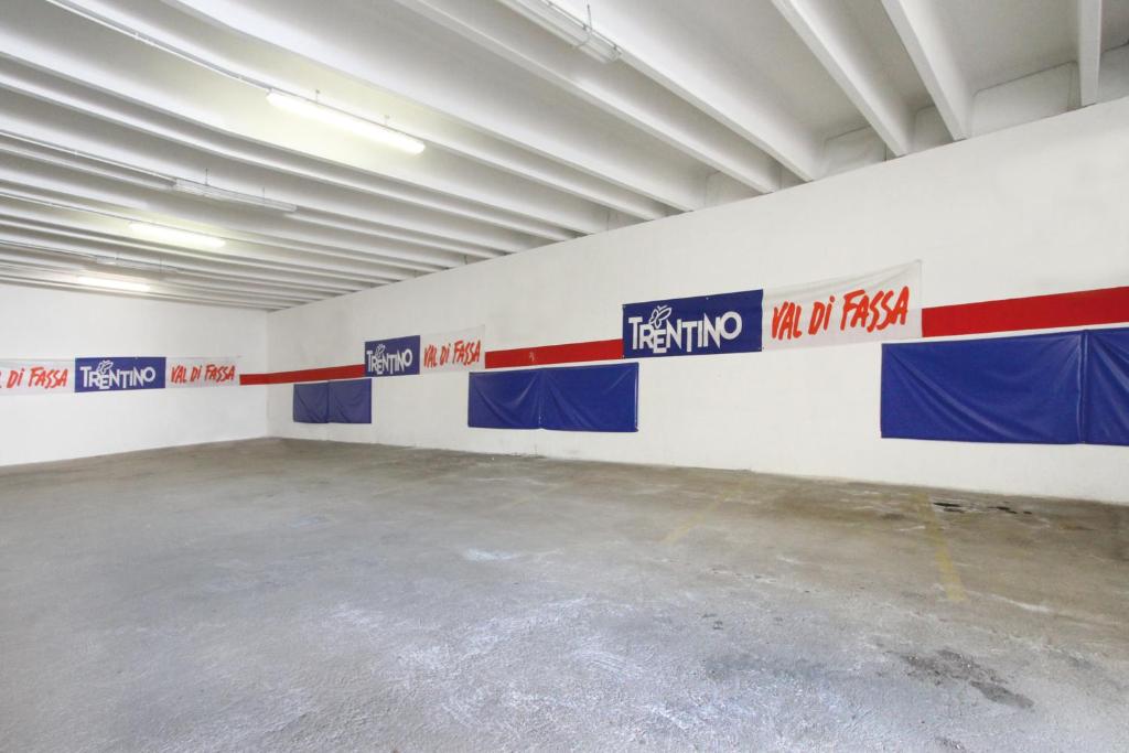 Tīra un plaša telpa ar baltiem sienām, kurās redzamas krāsainas reklāmas no Trentino un Val di Fassa. Grīdā redzama pelēka betona virsma ar dažām marķētām stāvvietām.