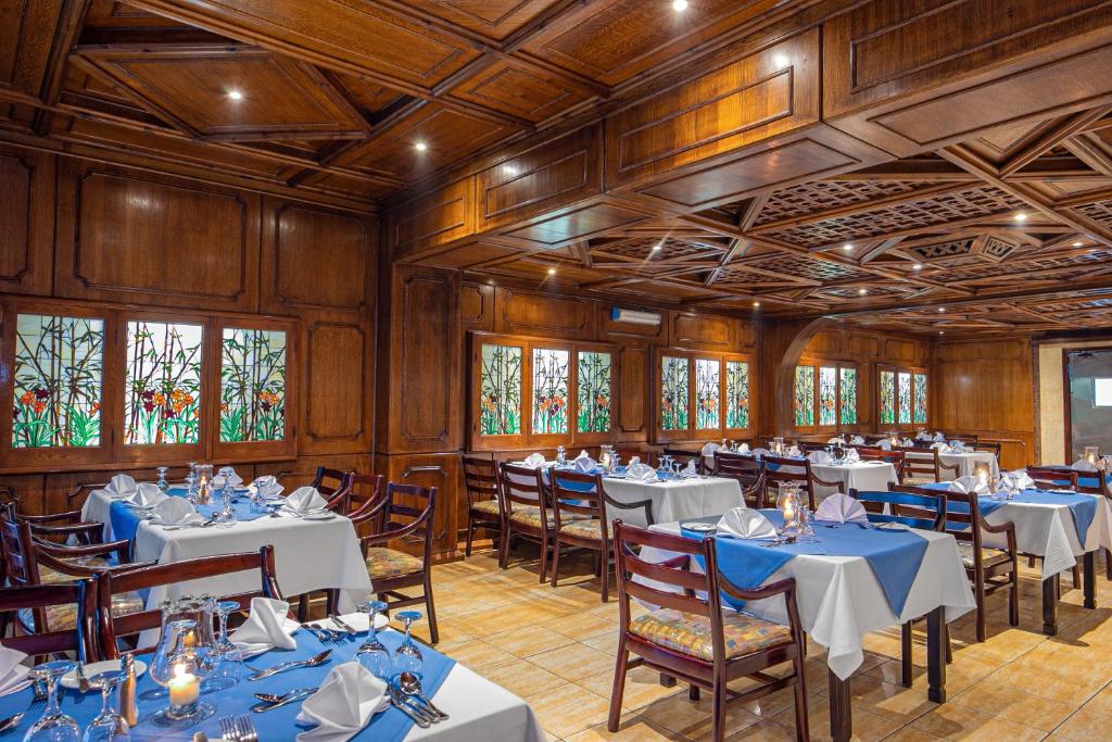 Elegants restorāna interjers ar koka apdari un krāsainiem stikla logiem, kas rada patīkamu atmosfēru. Galdi ir klāti ar baltām un ziliem galdautiem, nodrošinot izsmalcinātu ēdināšanas pieredzi.