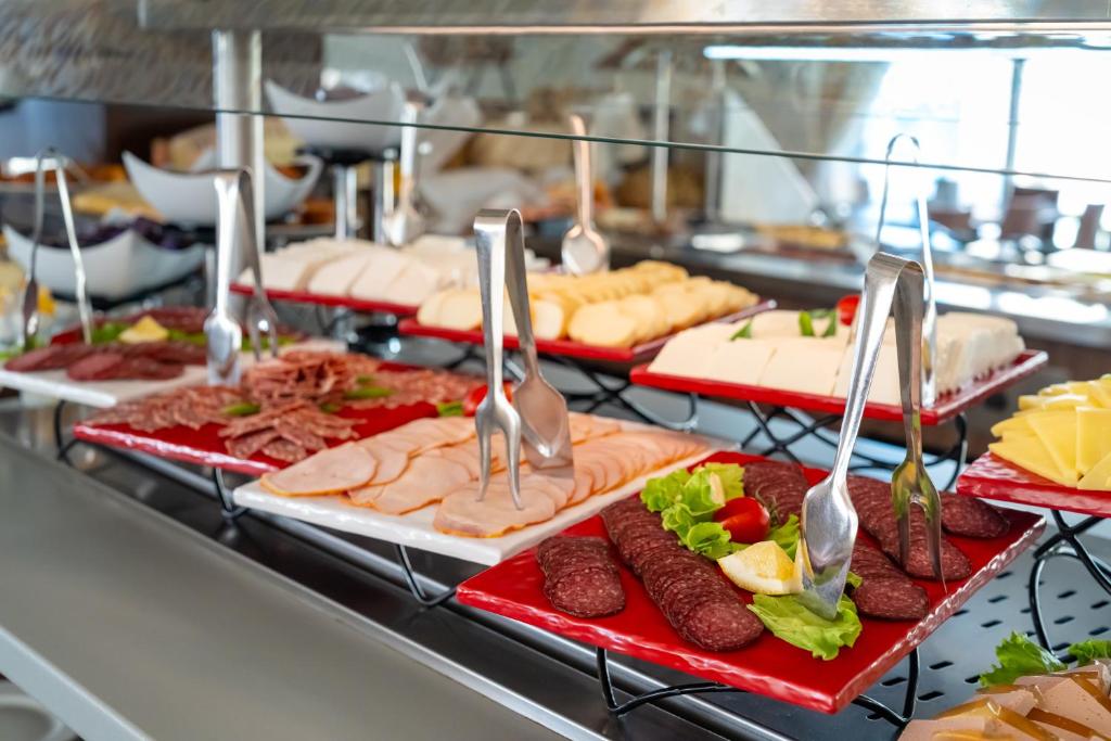 Pusdienu bufete ar dažādām gaļas un sieru šķēlēm, sakārtotām uz sarkaniem šķīvjiem, papildināta ar zaļumiem un tomātiem.