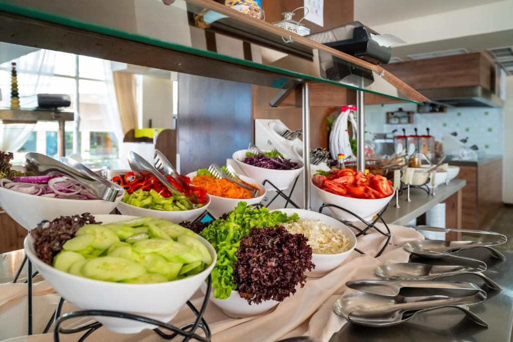 Salātu bufete ar dažādiem dārzeņiem, tostarp gurķiem, tomātiem, papriku un sīpoliem, pasniegta baltos traukos uz stilīgas galda.