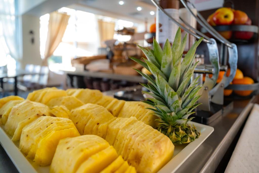 Svaigi sagriezta ananāsu šķēles uz balta šķīvja, blakus vesels ananāss, fons ar augļu groziem un saulainu interjeru.