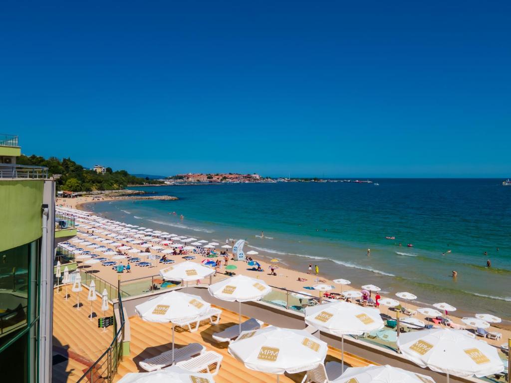 Pludmales skats ar baltām saules parasolēm un skaidru zilo ūdeni, kas piepildīts ar peldētājiem. Aiz pludmales redzama zaļa zeme un skaidrs debesis.