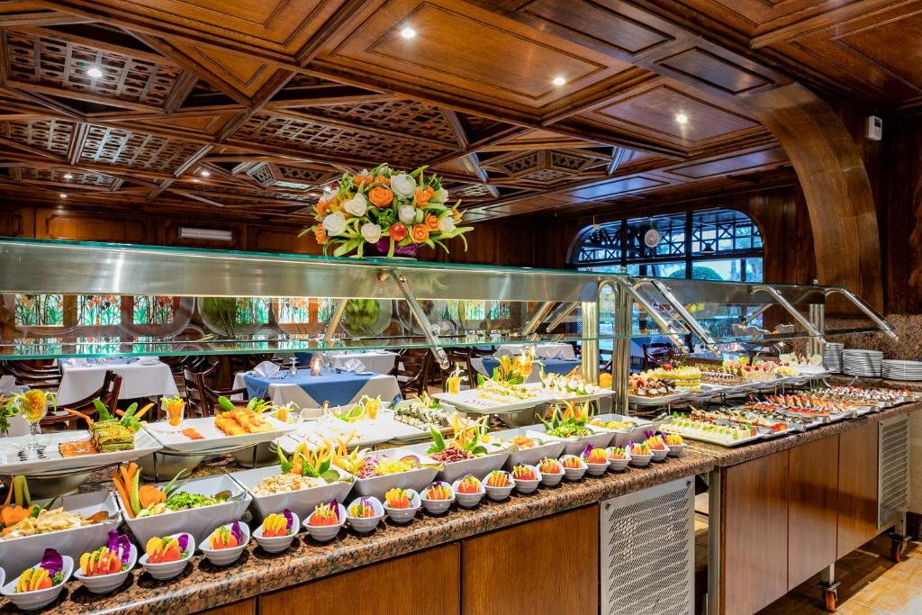Eleganti iekārtota restorāna bufete ar dažādiem kārumiem, tostarp dārzeņiem, augļiem un uzkodām, kas izkārtotas uz marmora galda, fona ar koka paneļiem un dekoratīvu ziedu kompozīciju.
