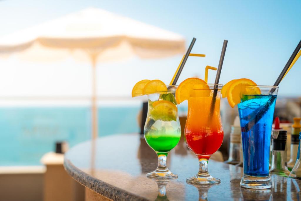 Krāsaini kokteiļi ar citrona šķēlītēm, novietoti uz bāra virsmas, fonā redzama jūra un saule. Ideāli piemēroti vasaras atpūtai.