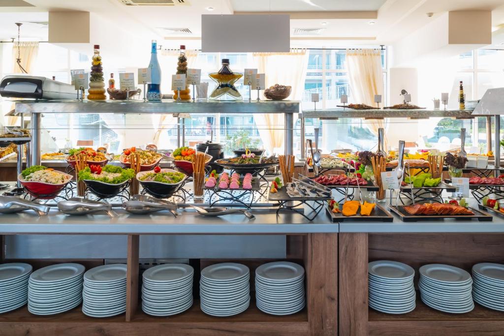 Bagātīgs bufete ar dažādiem ēdieniem, tostarp salātiem, gaļām un desertiem, izvietota pie elegantas viesnīcas. Galda apakšā redzami balti šķīvji.