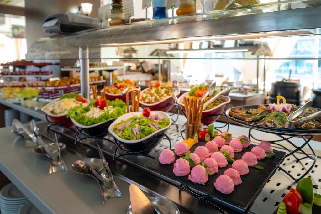 Bagātīgs bufetes galds ar dažādām salātiem, uzkodām un desertiem, tostarp rozā saldējumu un dārzeņiem. Galds ir dekorēts ar svaigiem dārzeņiem un garšvielām.