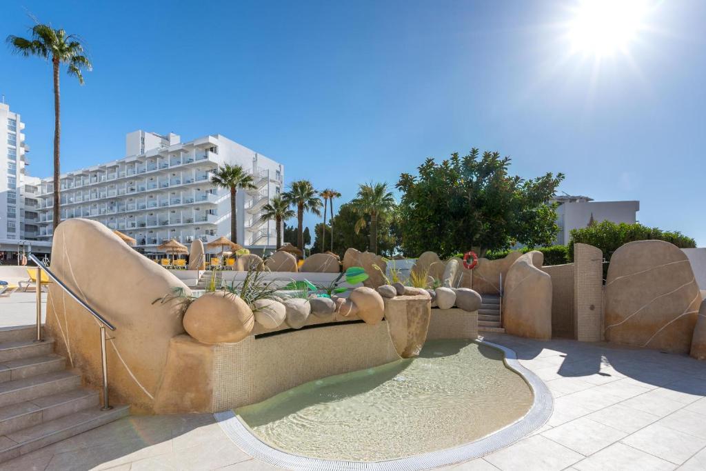 Vasaras brīvdienu komplekss ar peldbaseinu, kas ieskauts ar dabīgiem akmeņiem un palmām, ideāli piemērots relaksācijai saulainā dienā.