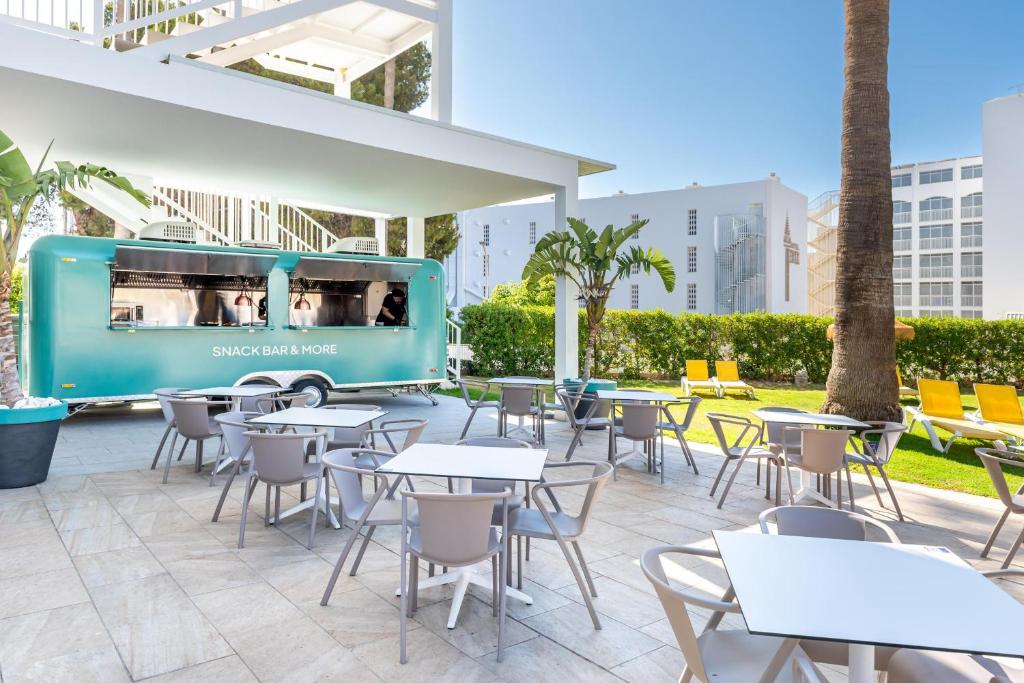 Āra kafejnīca ar modernu ēdienu kamioniņu, kurā redzami galdi un krēsli, kā arī zaļš zālienas fons un palmas. Ideāla vieta relaksācijai un uzkodām.