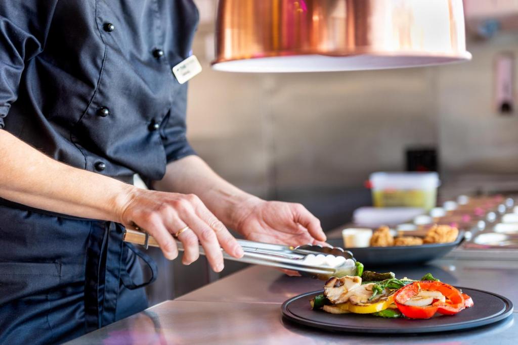 Šefpavārs gatavo maltīti, izmantojot knaibles, lai pasniegtu dārzeņus, kamēr strādā modernā virtuvē. Uz galda redzami dažādi ēdieni un rīki.