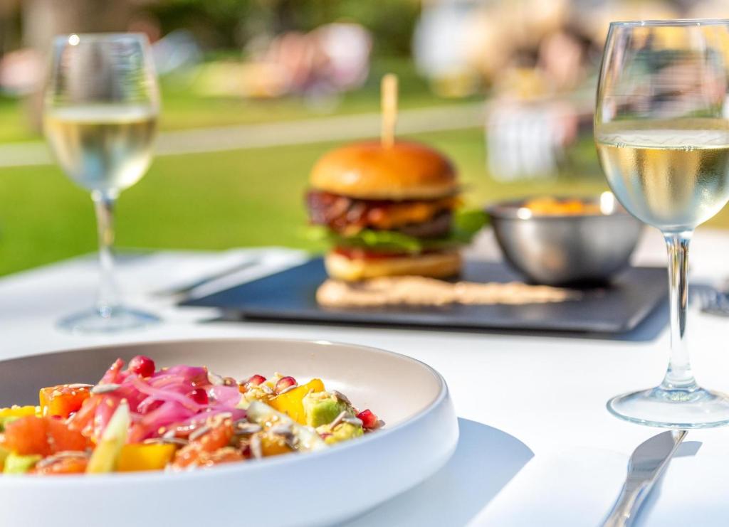 Svaigu dārzeņu salāti ar avokado un granātābolu sēklām, blakus sulīgs burgeris un glāze balto vīna, radot ideālu ēdiena baudīšanas mirkli brīvā dabā.