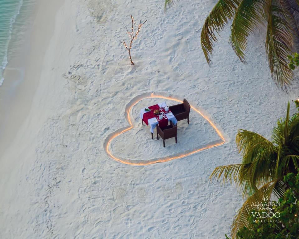 Romantisks galda iestatījums sirdī veidotā smiltīs pludmalē, ar dekoratīvām gaismām un tropiskām palmām apkārt. Ideāla vieta romantiskām vakariņām Maldivās.