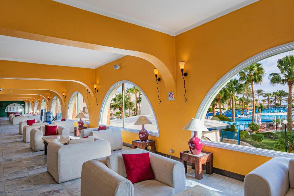 Elegants viesnīcas interjers ar dzeltenām sienām un plašiem logiem, redzama peldbaseina un palmu ainava. Mīkstās mēbeles ar sarkaniem spilveniem veido mājīgu atmosfēru.