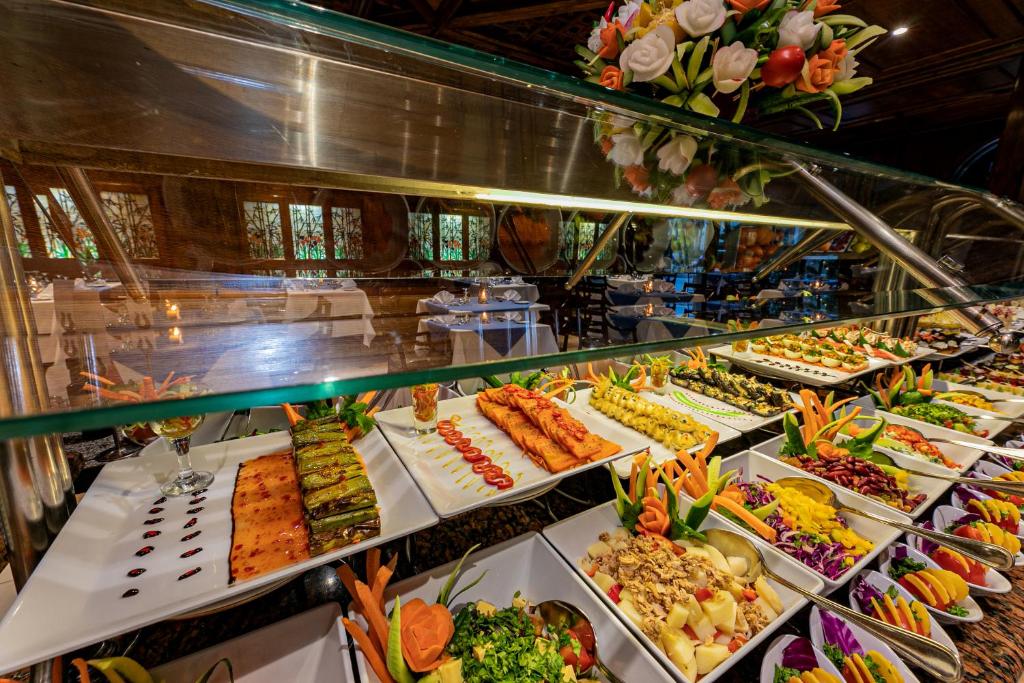 Bagātīgs bufete ar dažādiem ēdieniem, tostarp dārzeņu salātiem, zivi un garnelēm, rotāta ar krāsainām dekorācijām. Restorāna iekšiene ar elegantām galdiem un gaismu rada patīkamu atmosfēru.