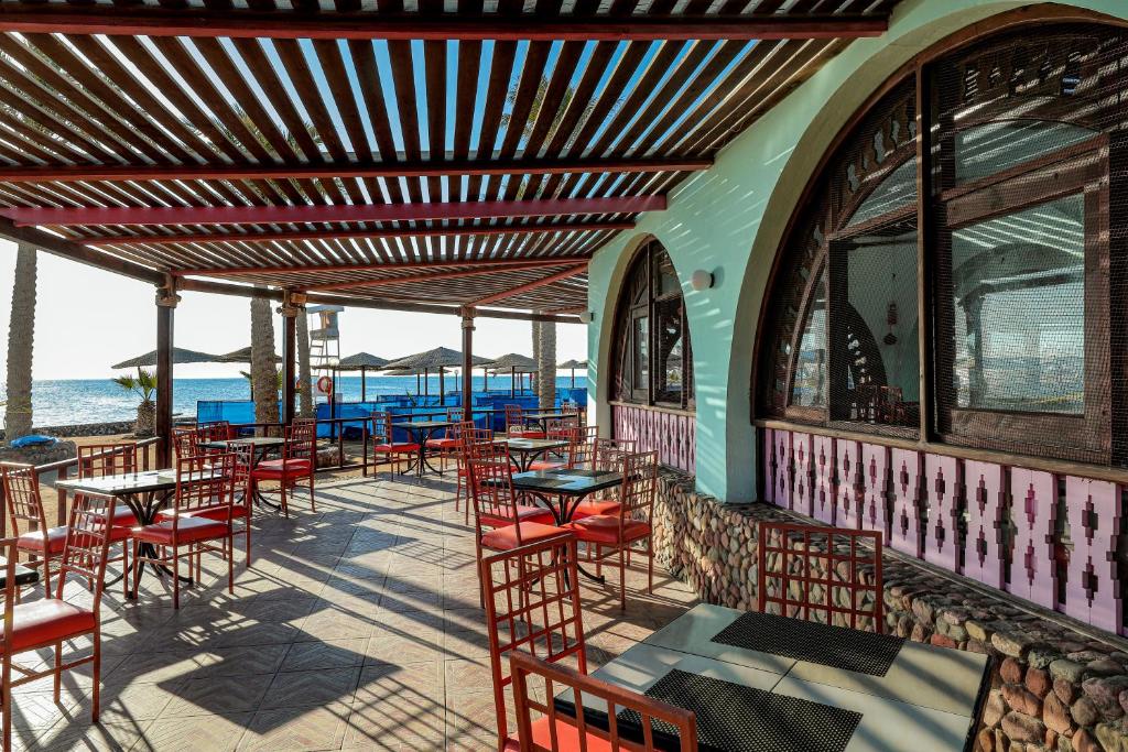 Atvērts restorāns ar terasi pie jūras, kurā ir sarkanas krēsli un galdi, zem koka nojumes. Aiz terases redzamas palmu un pludmales ainavas.
