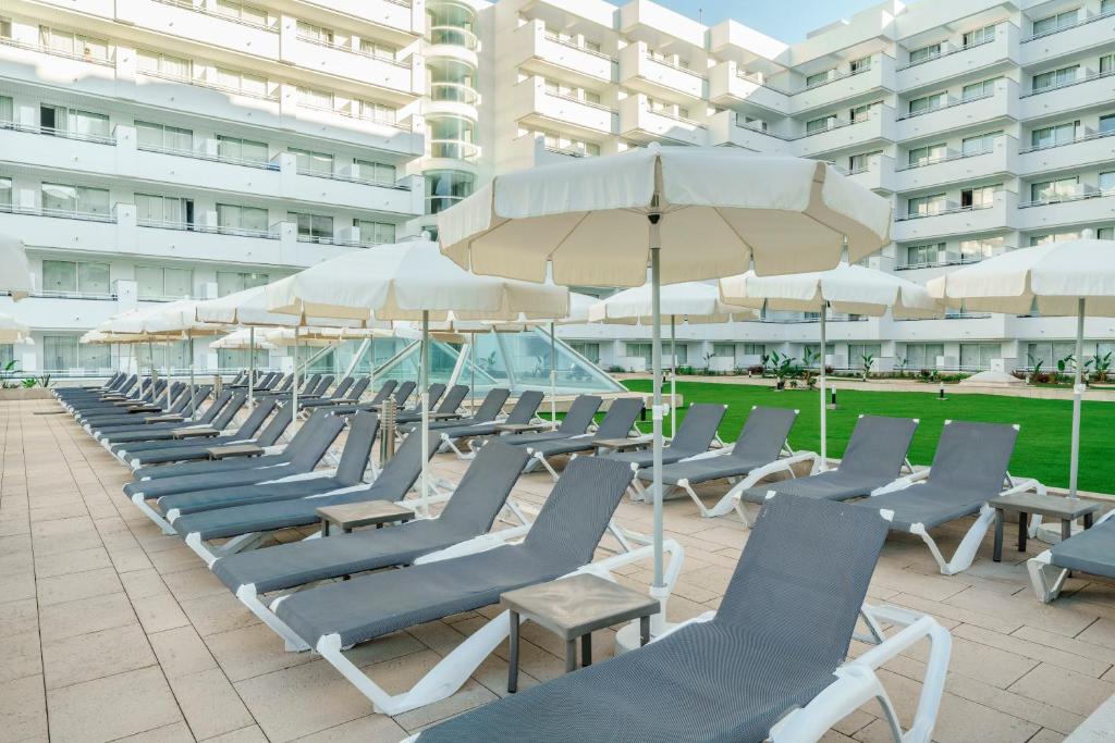 Baseina zona ar sauļošanās krēsliem un saulessargiem, kas izvietoti blakus modernai viesnīcai. Zaļš zālienis papildina atpūtas vidi.