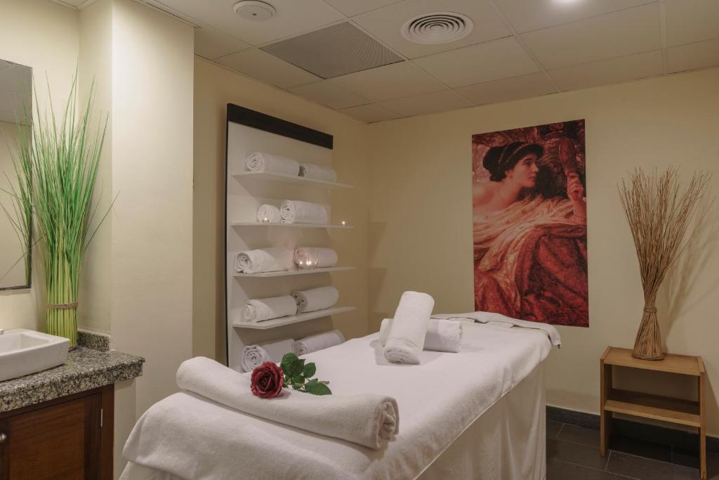 Relaksējoša spa telpa ar masāžas galdu, uz kura ir dvieļi un sarkana roze, kā arī dekoratīvi elementi, piemēram, augi un gaismiņas. Sienā ir gleznas reprodukcija, kas piešķir telpai mājīgu atmosfēru.