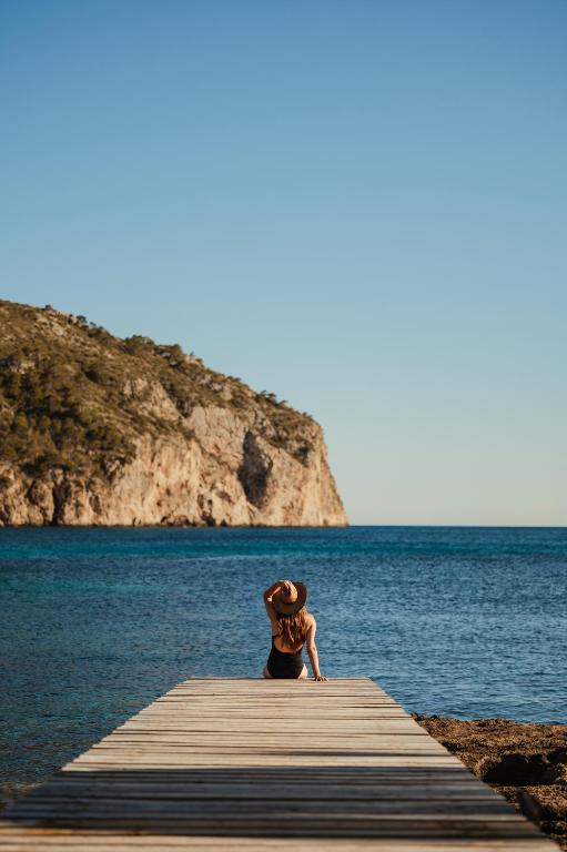 Sieviete, kas sēž uz koka mola pie jūras, ar skatu uz klintīm un mierīgu ūdeni. Ideāla aina relaksācijai un atpūtai pie dabas.