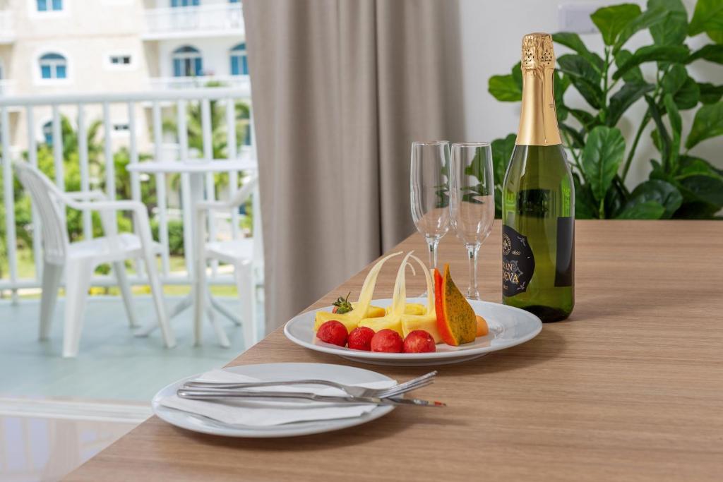 Svaigu augļu plate ar šampanieša pudeli un glāzēm, izvietota uz koka galda. Fons ar balkonu un zaļiem augiem rada relaksējošu atmosfēru.