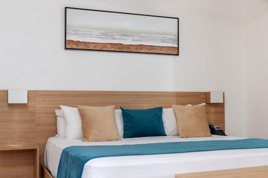 Mūsdienīga guļamistaba ar koka gultu un dažādiem spilveniem, kas papildināta ar ainavu gleznu uz sienas. Gultas veļa ir vienkārša, bet stilīga, radot relaksējošu atmosfēru.