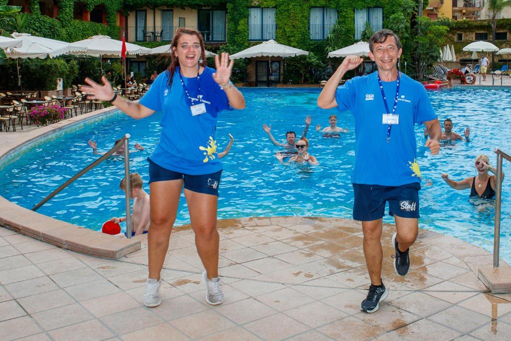 Divi cilvēki jautri dejo pie baseina, kamēr aizmugurē redzams grupiņa peldētāju. Abiem ir zila T-kreklu, un viņi izskatās priecīgi, piedaloties aktivitātēs. Pilsētas ainava un zaļi augi veido patīkamu fonu.