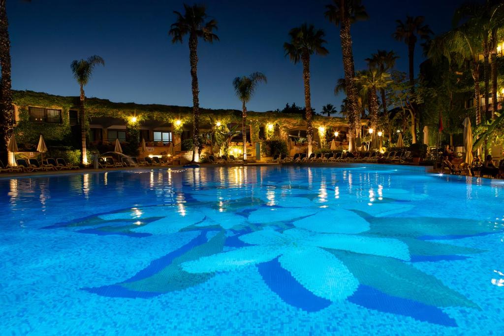 Nakts ainava ar apgaismotu baseinu, kurā redzama ziedu mozaīka un apkārt esošas palmas. Mierīga atmosfēra un siltas gaismas rada relaksējošu noskaņu.