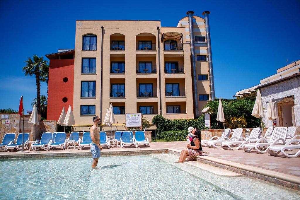 Baseins ar atpūtas zonu, kurā cilvēki bauda sauli, fons veido moderna viesnīca un zils debesis.