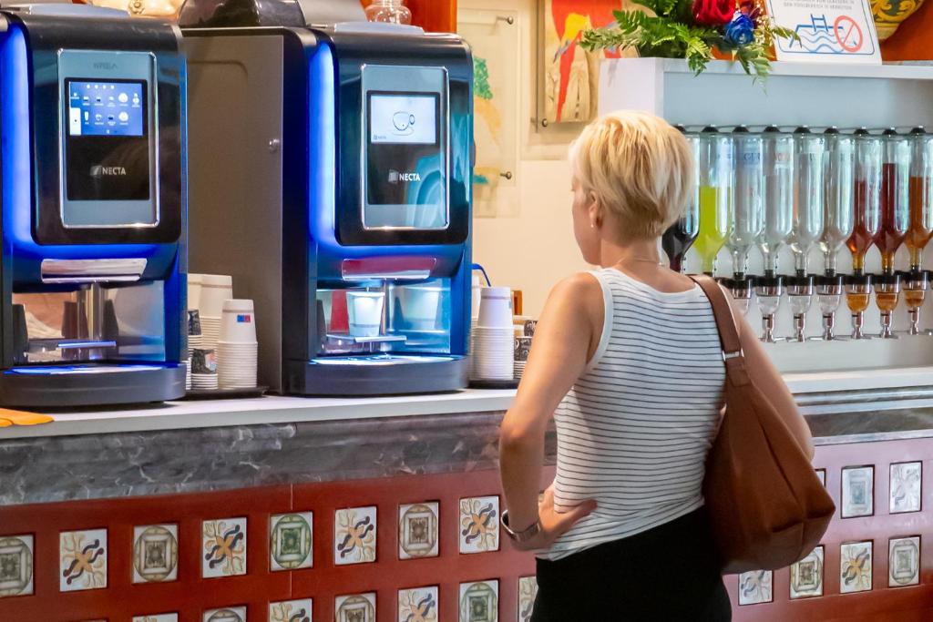 Sieviete ar īsiem matiem skatās uz divām kafijas automātām, kas atrodas kafejnīcā. Fona sienā redzami dažādi dzērieni un mākslas darbi.