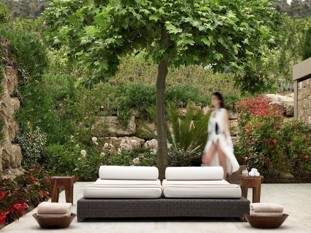 Mūsdienīgs dārza stūrītis ar ērtu dīvānu un dekoratīvām augu kompozīcijām. Fonā redzama persona balta apģērbā, kas pastaigājas pa dārzu.
