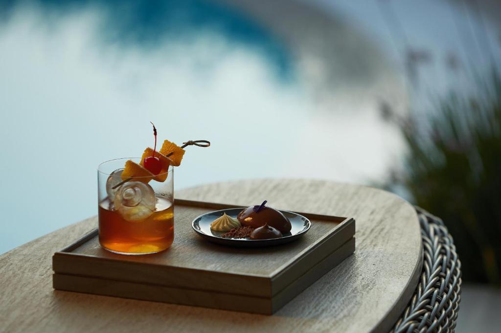 Kokteilis ar apelsīnu un ķiršu rotājumu uz koka trauka, blakus neliela šķīvis ar saldumiem, fons ar baseinu un zaļumiem.