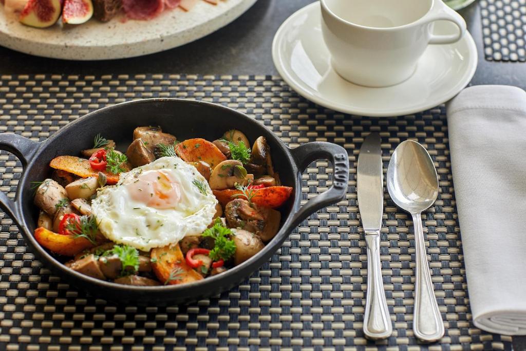 Sautēti dārzeņi ar olu un sēnēm, pasniegti stilīgā pannā, blakus balta kafijas tasīte un piederumi. Ideāls brokastu vai pusdienu ēdiens.