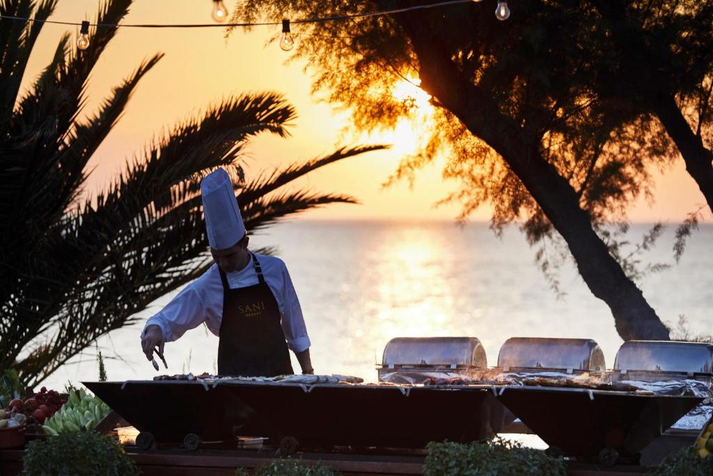Šefpavārs gatavo ēdienus pie grila saulrieta laikā, ar jūru un palmām fona. Gaismas virtenes piešķir burvīgu atmosfēru.