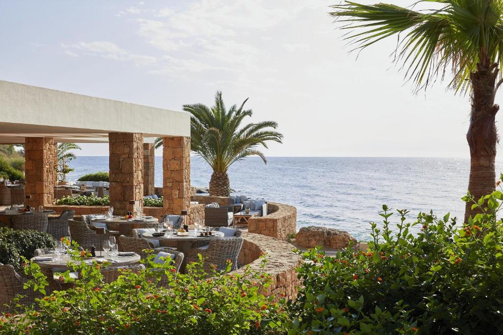 Jūras krastā esošā restorāna terase ar ēdamgaldiem un palmām, kas rada mierīgu un relaksējošu atmosfēru. Ideāla vieta gastronomiskām baudām pie jūras.