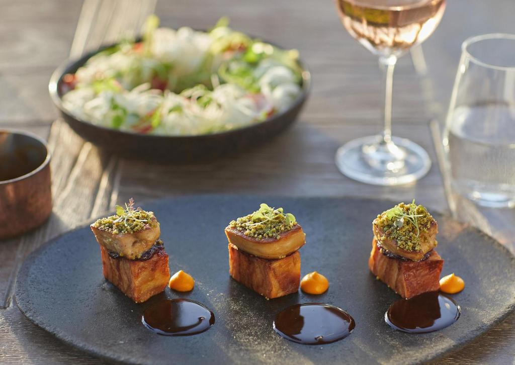 Elegants ēdiens, kurā redzami trīs mazi gaļas gabaliņi ar garšaugiem un mērci, pasniegti uz melnas šķīvja. Fona salātu trauks un dzēriens papildina gastronomisko pieredzi.