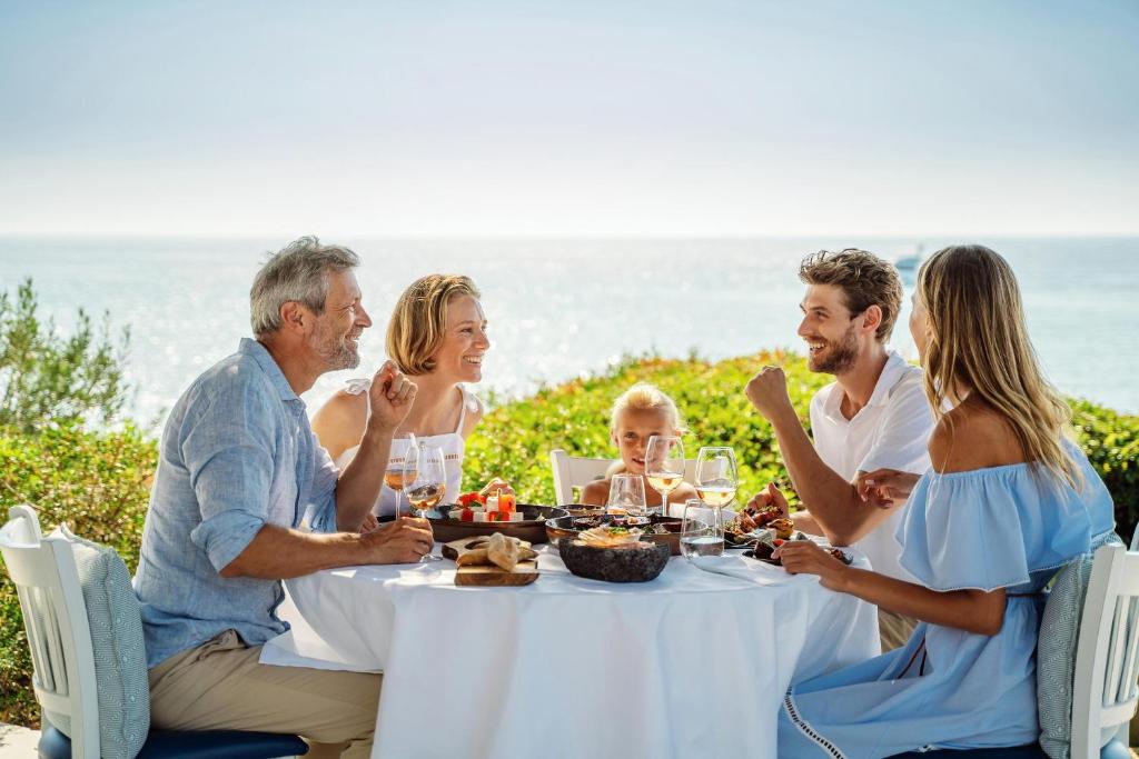 Ģimene baudījusi maltīti pie galda ar skatu uz jūru, svinot laimīgas brīvdienas un daloties stāstos. Galda centrā ir dažādi ēdieni un dzērieni.