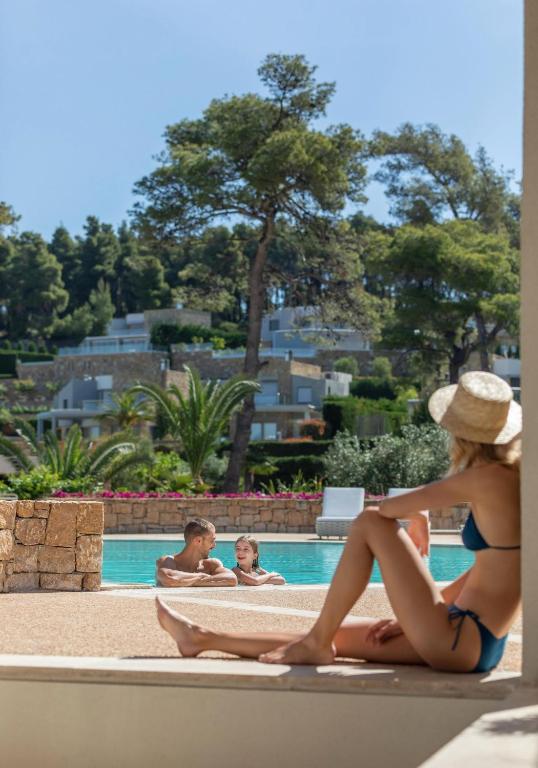 Peldbaseinsā atpūšas sieviete ar vāciņu, kamēr divi bērni priecīgi spēlējas ūdenī. Apkārt ir zaļa daba un moderni mājokļi.