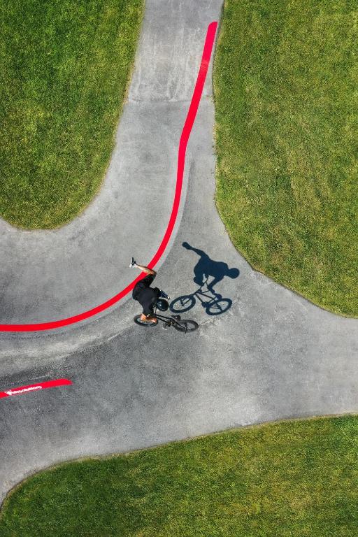 Velosipēdists brauc pa ceļu, kas iezīmēts ar sarkanu līniju, ar zaļām zālēm apkārt. Perspektīva no augšas uzsver dinamiku un kustību.