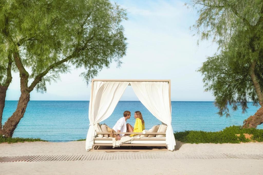 Romantiska pāris atpūšas pludmalē zem baldahīna ar skatu uz jūru, ko ieskauj zaļi koki.