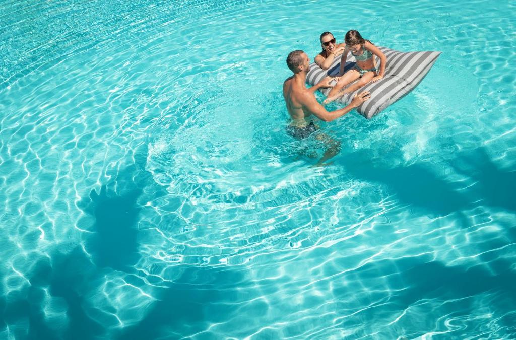 Ģimenes laiks pie baseina, kur pieaugušie un bērns atpūšas uz pludiņa, bauda sauli un ūdeni.