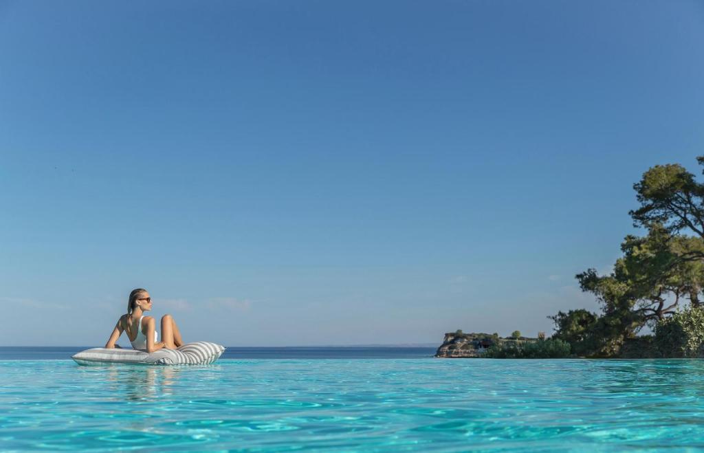 Sieviete atpūšas uz peldēšanas spilvena kristāldzidra ūdens baseinā, ar jūru un debesīm fonā. Ideāla ainava relaksācijai un brīvā laika pavadīšanai.