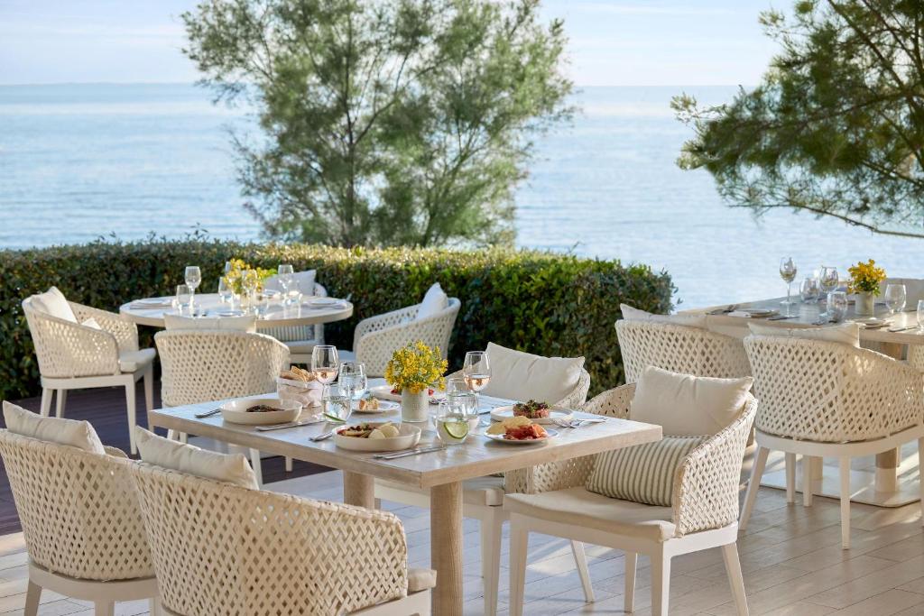Elegants restorāns ar skatu uz jūru, kur baltas mēbeles un ziedu kompozīcijas radījušas patīkamu atmosfēru. Galdi ir klāti ar gardām maltītēm un vīnu, piedāvājot relaksējošu ēšanas pieredzi.