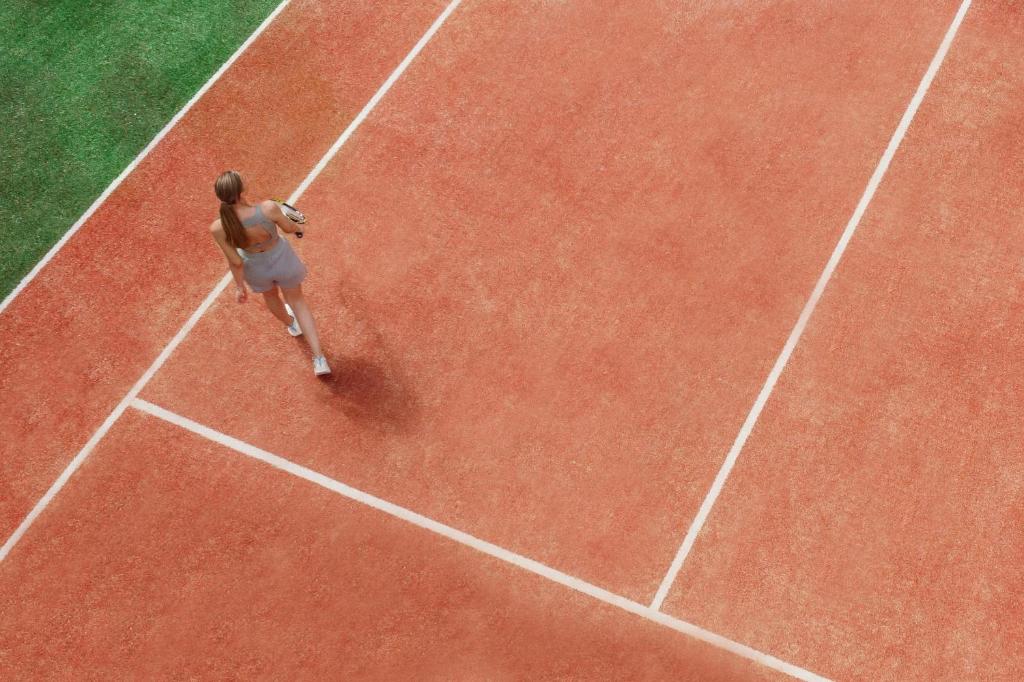 Sieviete, tērpta sportiskā apģērbā, pastaigā pa tenisa laukumu ar oranžu segumu, turot raketi. Fons ir zaļš zālājs.